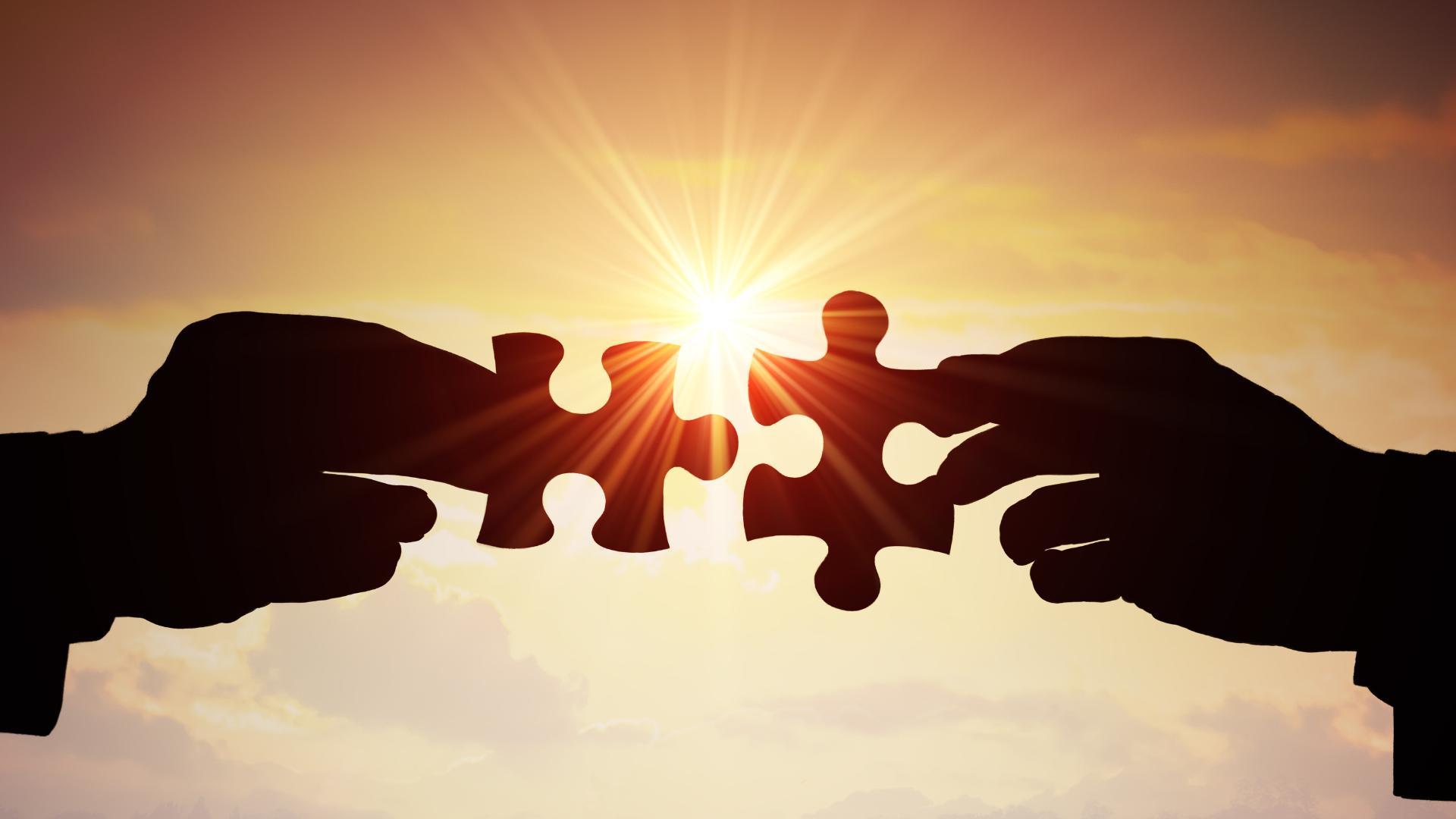 Deux mains rapprochant des pièces de puzzle devant un coucher de soleil, symbolisant la collaboration, l'unité et la résolution de problèmes.