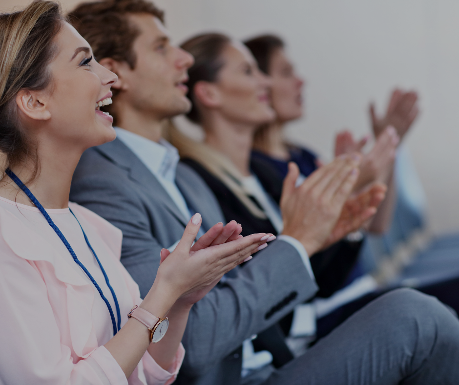 Un groupe de professionnels assis en rangée lors d'un événement, applaudissant avec enthousiasme. Ils sont concentrés sur ce qui semble être une présentation ou une performance.