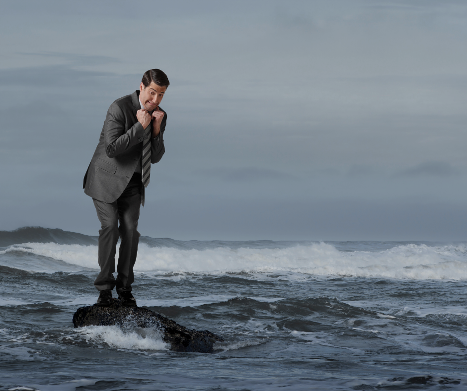 Homme en costume debout sur un rocher isolé entouré par des vagues agitées, exprimant l'inquiétude ou la peur, symbolisant les défis, l'isolement ou la gestion du stress en milieu professionnel.